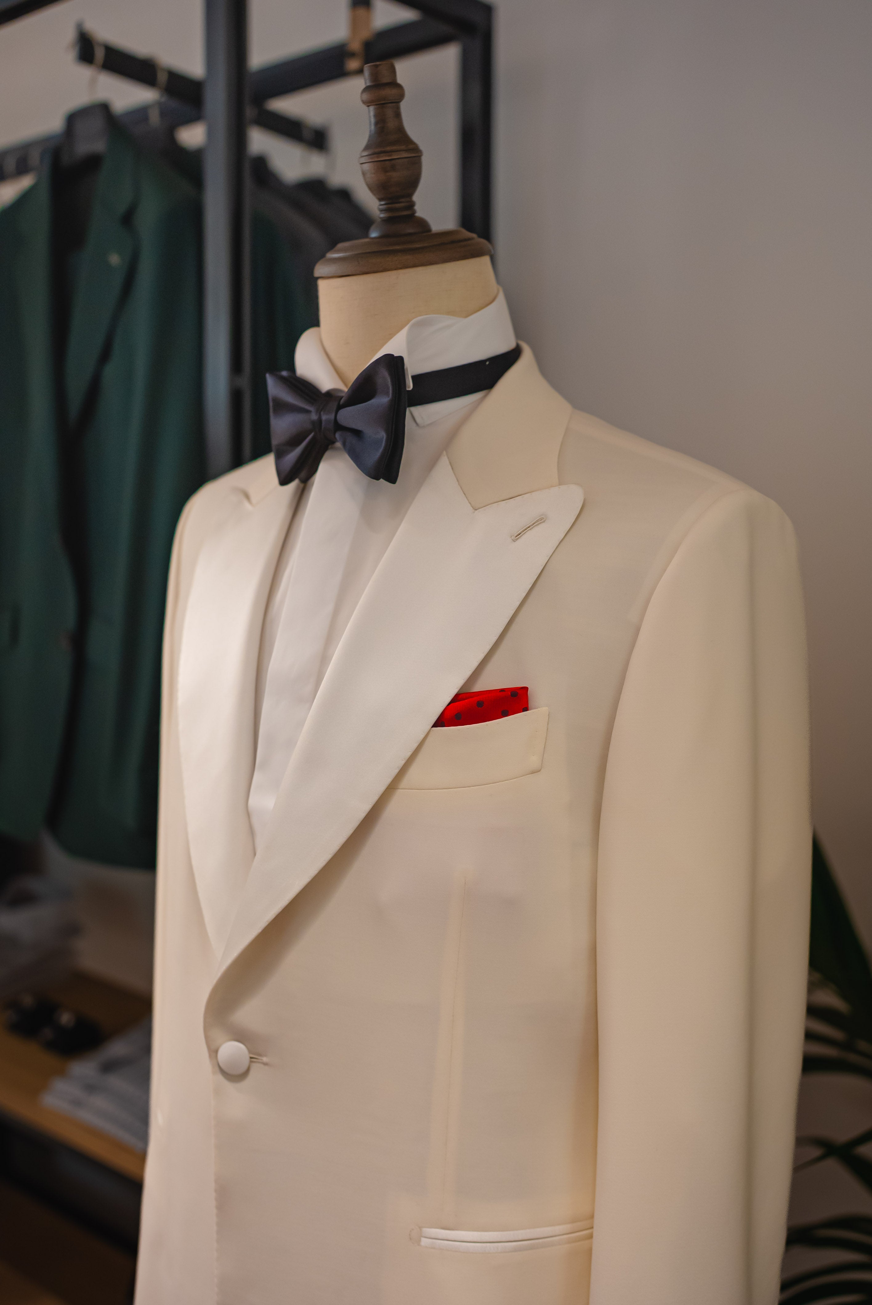 White suit jacket with a black bow tie and red pocket square on a mannequin, with a blurred background of clothing racks. - Sartoria Dei Duchi-Atri