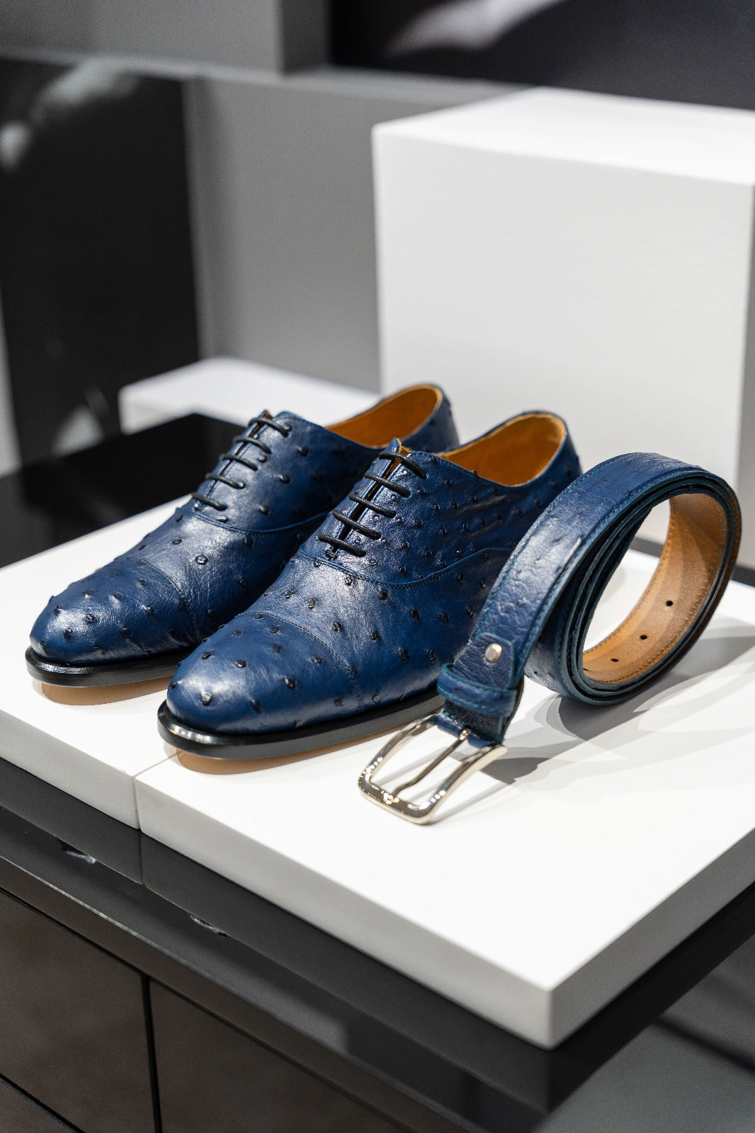 Blue dress shoes with a matching belt on a white platform. - Blue leather belt with a gold buckle on a white surface. - Sartoria Dei Duchi-Atri